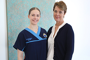 Two women stand in front of a piece of artwork. The woman on the left wears a nurses uniform.