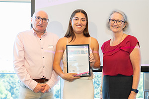 Monique Carbone stands between a man and woman. She holds a WANMEA 2025 finalist certificate.