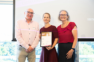 Adele King stands between a man and woman. She holds a WANMEA 2025 finalist certificate.