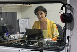 A woman sits in a sealed off booth looking at a computer. On this side of the window hangs a pair of earphones