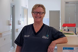 A woman wearing a Fiona Stanley Hospital uniform that reads Clinical Nurse Consultant leans against a counter