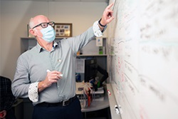 An older man wearing a surgical mask writes on a whiteboard.