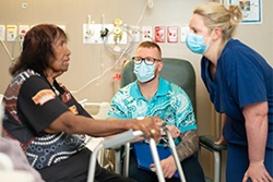 An Aboriginal woman sits on the side of a bed holding the top of a walking frame as she talks with a male and female health professional wearing surgical masks.