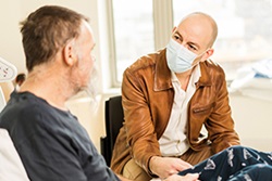 A man wearing a surgical mark sits talking to a man sitting in a hospital bed.