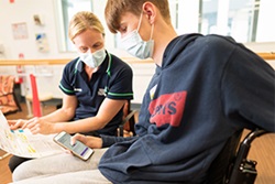 A female health professional talks with a young man who is seated in a wheelchair. He is holding smartphone.