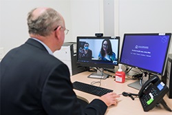 A man sitting in front of two computer screen during an online meeting. Two people can be seen on one of the screens.