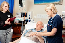 A nurse treats an older man who is laying in a bed. Another woman stands beside the bed writing on a clipboard.