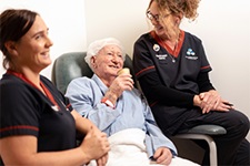 Two female nurses sit beside an older man who is seated and sipping from a small cup