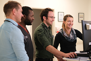 Three men and a woman look at a computer screen. One of the men wears a shirt that reads 'Registered nurse'