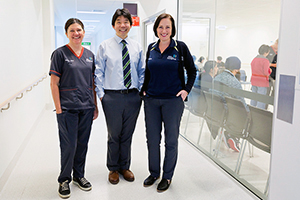 A male and two female health professionals stand in a hospital hallway. Patients can be seen waiting in an area behind them.