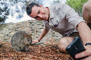 A young man reclines on the ground and leans back to take a photo with a nearby quokka