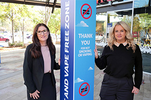 Two women stand beside vertical smoke and vape free signage  
