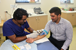 A male health professional inserts a peripheral intravenous catheter (PIVC,or cannula) into the right forearm of another man seated beside him