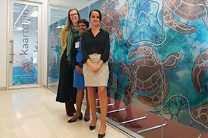 Three women stand in front a window panels featuring Aboriginal artwork turtles. On the door behind them is the word 'Kaartdijin'.