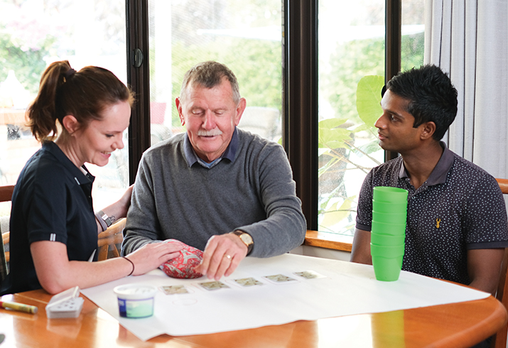 An older man sits at a dining table in a home setting between a female and male therapist