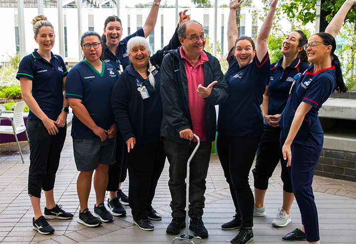 An older man using a walking stick for support stands with a group of health professionals