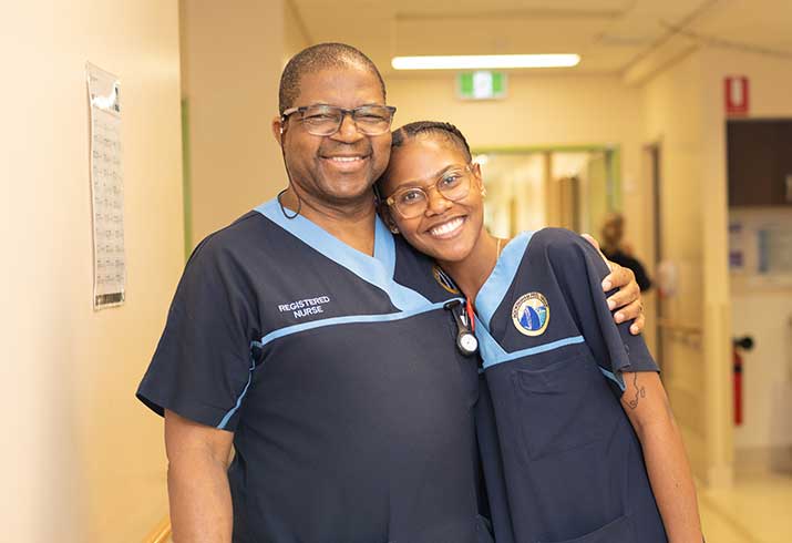 A male nurse and female nurse stand in a hospital hallway.