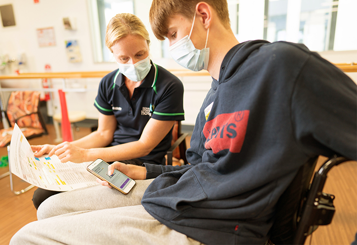 A female health professional talks with a young man who is seated in a wheelchair. He is holding smartphone.