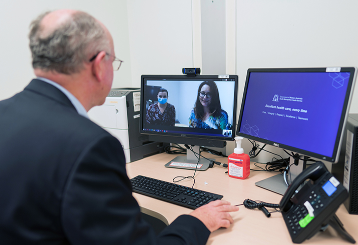 A man sitting in front of two computer screen during an online meeting. Two people can be seen on one of the screens.