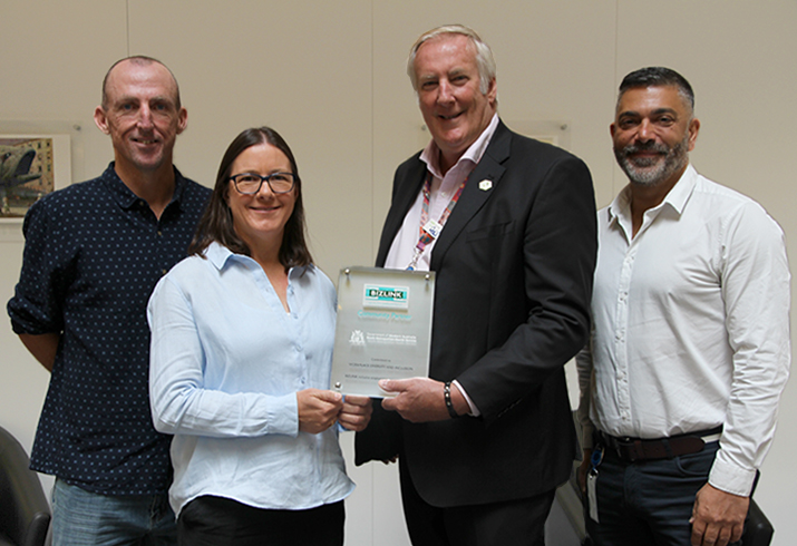 Three men and one woman stand together. One man and the woman are jointly holding a framed certificate.