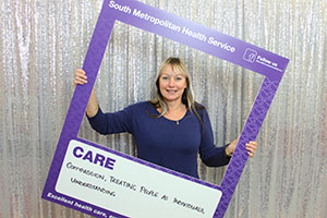 A woman stands inside a frame that reads 'South Metropolitan health Service: Care'. A handwritten message on the board reads 'Compassion. Treating people as individuals. Understanding.'