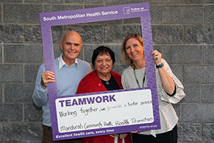 A man and two woman stands inside a frame that reads 'South Metropolitan Health Service: Teamwork.'A handwritten message on the board reads 'Working together to provide a better service – Mandurah Community Health, Health Promotion.' 