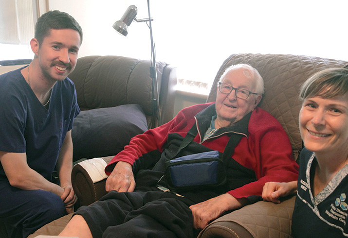 A male doctor in training and a female nurse sit beside an older male Home Hospital patient