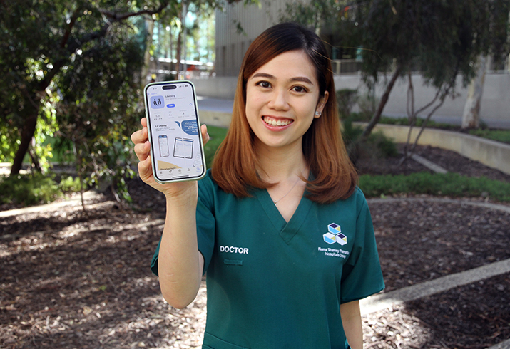 A young woman wearing Fiona Stanley Fremantle Hospitals Group scrubs holds a mobile phone with an open app on the screen.